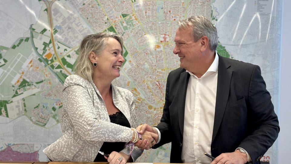 Joziene van de Linde, Managing Director bij VDL De Meeuw, en Carlo Schreuder, Directeur Ruimtelijk Economische Opgave bij gemeente Eindhoven, schudden elkaar de hand bij de feestelijke ondertekening van een samenwerkingsovereenkomst voor flexibele huisvesting in Eindhoven. Op de achtergrond is een stadsplattegrond van Eindhoven zichtbaar.