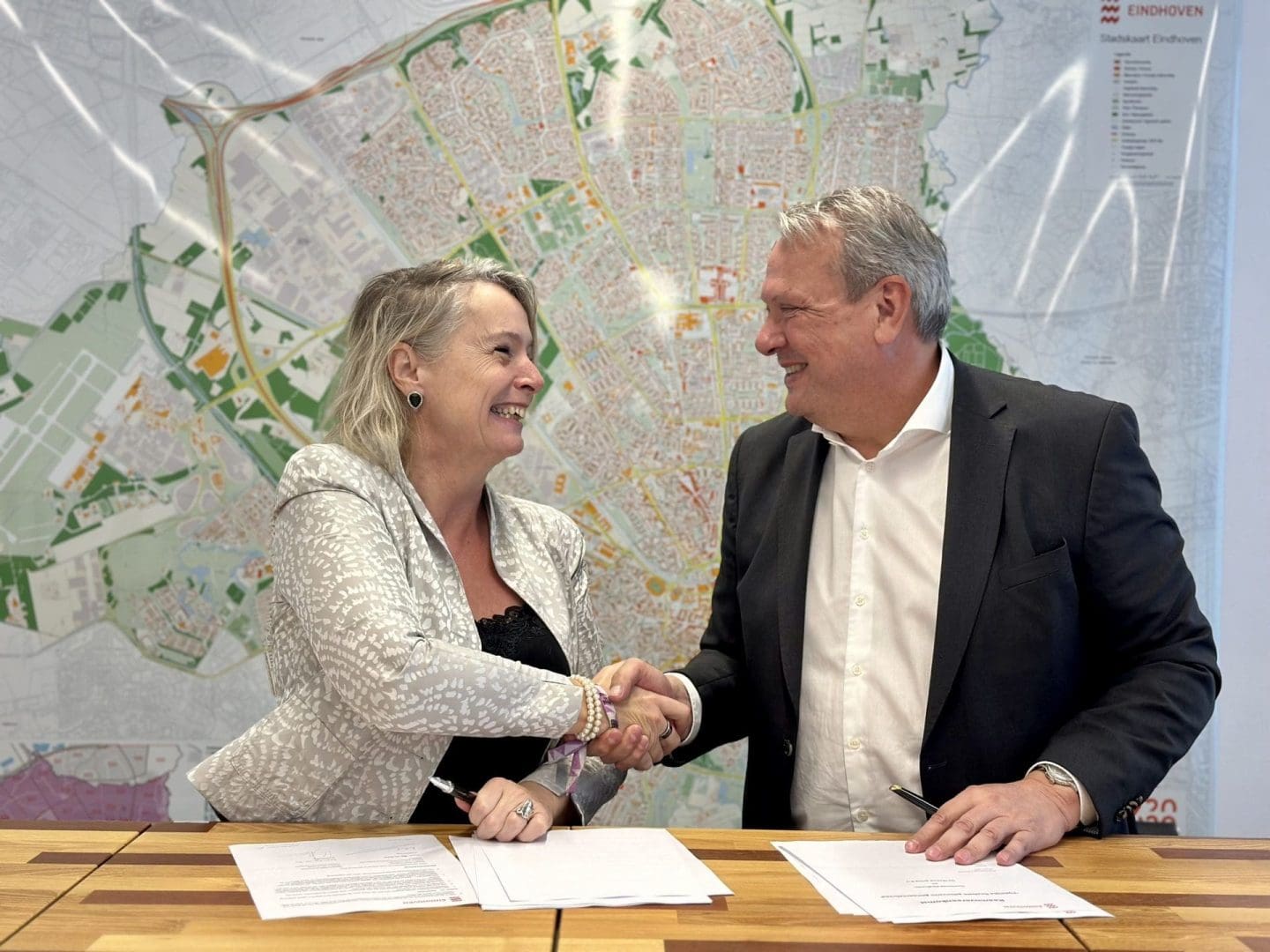 Joziene van de Linde, Managing Director bij VDL De Meeuw, en Carlo Schreuder, Directeur Ruimtelijk Economische Opgave bij gemeente Eindhoven, schudden elkaar de hand bij de feestelijke ondertekening van een samenwerkingsovereenkomst voor flexibele huisvesting in Eindhoven. Op de achtergrond is een stadsplattegrond van Eindhoven zichtbaar.
